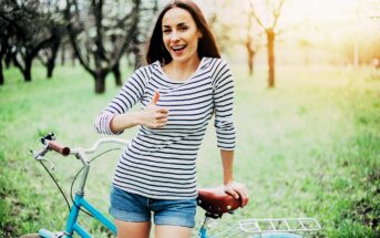 A woman in a striped shirt and denim shorts stands next to a bicycle in a grassy park, smiling and giving a thumbs-up. The background features trees and sunlight filtering through the branches.