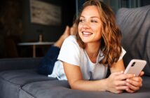 A woman with shoulder-length curly hair is lying on a gray couch, smiling while holding a smartphone. She is wearing a white t-shirt and jeans, and the background shows a blurred living room setting.