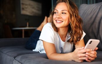 A woman with shoulder-length curly hair is lying on a gray couch, smiling while holding a smartphone. She is wearing a white t-shirt and jeans, and the background shows a blurred living room setting.