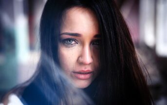 A person with long dark hair and striking blue eyes looks directly at the camera. There are streaks of makeup running down their cheeks, suggesting tears. The background is blurred, emphasizing their emotional expression.