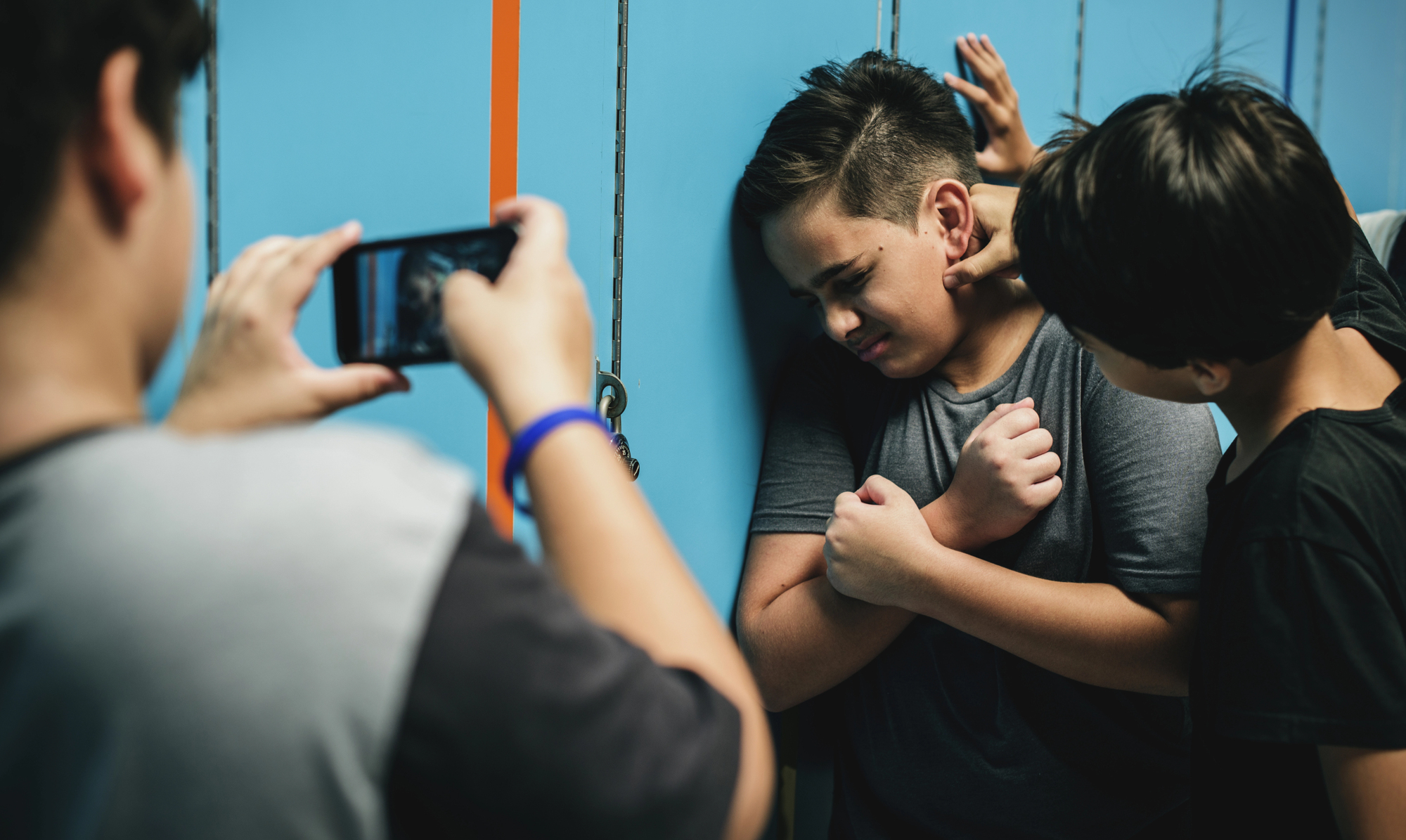 A boy is being bullied by another boy in front of a set of blue lockers. A third person records the incident with a smartphone from the side. The bullied boy looks distressed and covers his face with his arms.