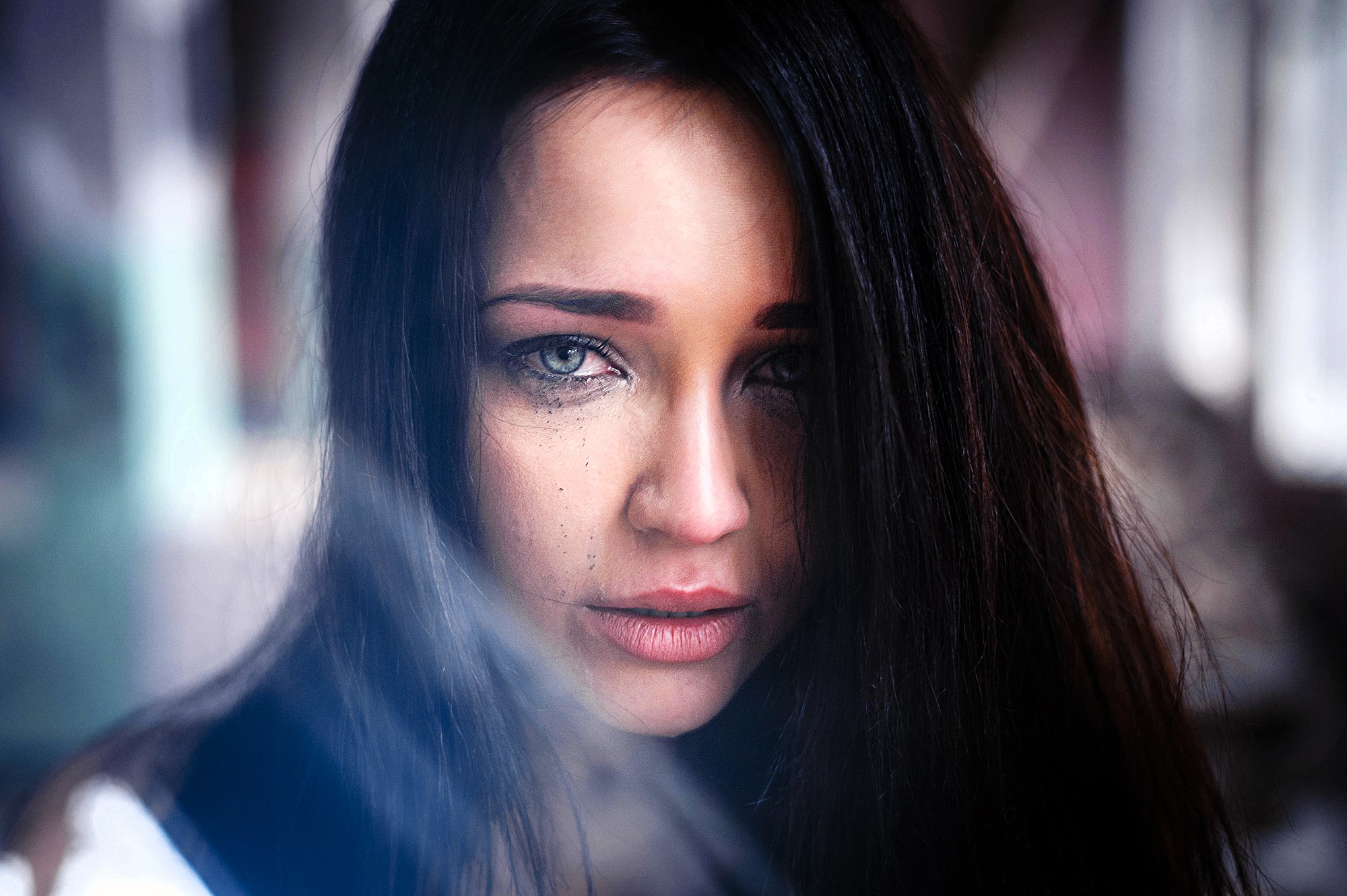 A person with long dark hair and striking blue eyes looks directly at the camera. There are streaks of makeup running down their cheeks, suggesting tears. The background is blurred, emphasizing their emotional expression.