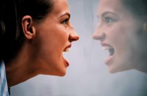 A woman with a tense expression and open mouth faces her reflection in a mirror. The reflection mirrors her intense expression, creating a symmetrical and emotional scene.