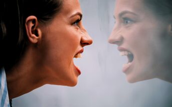 A woman with a tense expression and open mouth faces her reflection in a mirror. The reflection mirrors her intense expression, creating a symmetrical and emotional scene.