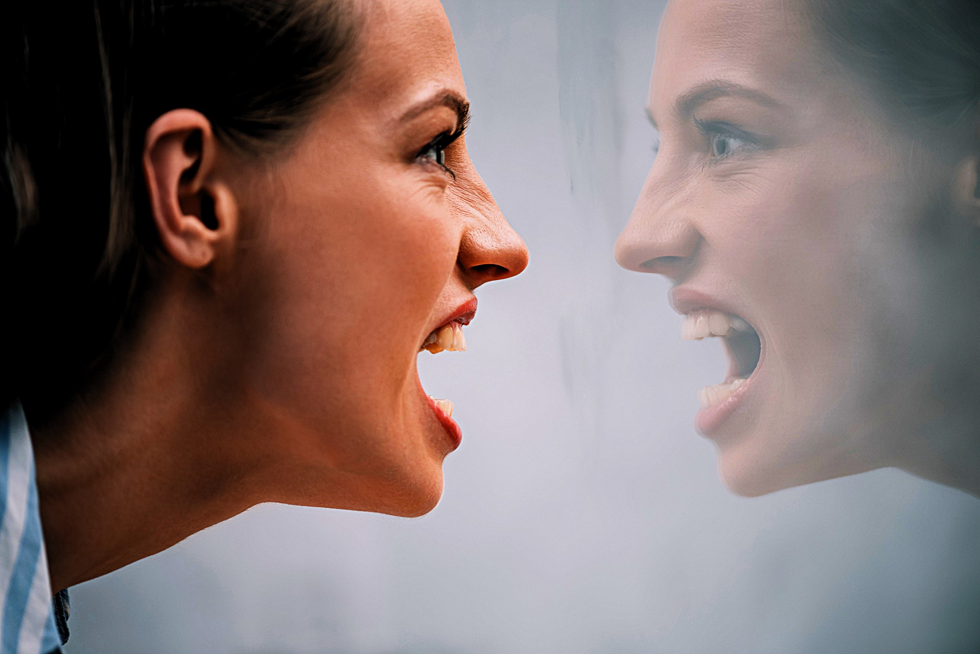 A woman with a tense expression and open mouth faces her reflection in a mirror. The reflection mirrors her intense expression, creating a symmetrical and emotional scene.