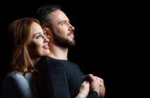 A couple stands against a black background, smiling and gazing upwards. The woman, with long brown hair, embraces the man from behind. Both wear long-sleeved shirts, hers light gray and his dark striped. They exude happiness and affection.