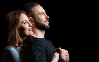 A couple stands against a black background, smiling and gazing upwards. The woman, with long brown hair, embraces the man from behind. Both wear long-sleeved shirts, hers light gray and his dark striped. They exude happiness and affection.