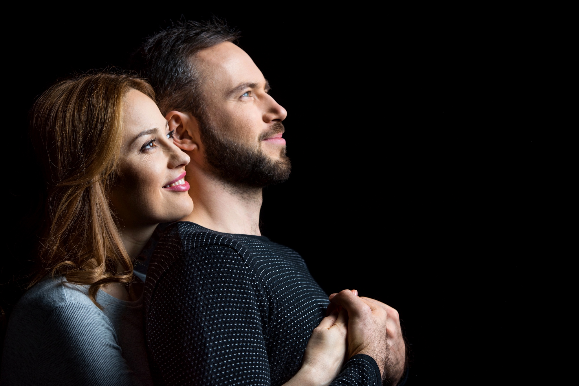 A couple stands against a black background, smiling and gazing upwards. The woman, with long brown hair, embraces the man from behind. Both wear long-sleeved shirts, hers light gray and his dark striped. They exude happiness and affection.