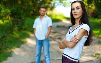 A woman with long dark hair stands in the foreground with arms crossed, wearing a white shirt, and looking at the camera. A man in a white shirt and blue jeans stands blurred in the background on a path surrounded by greenery.