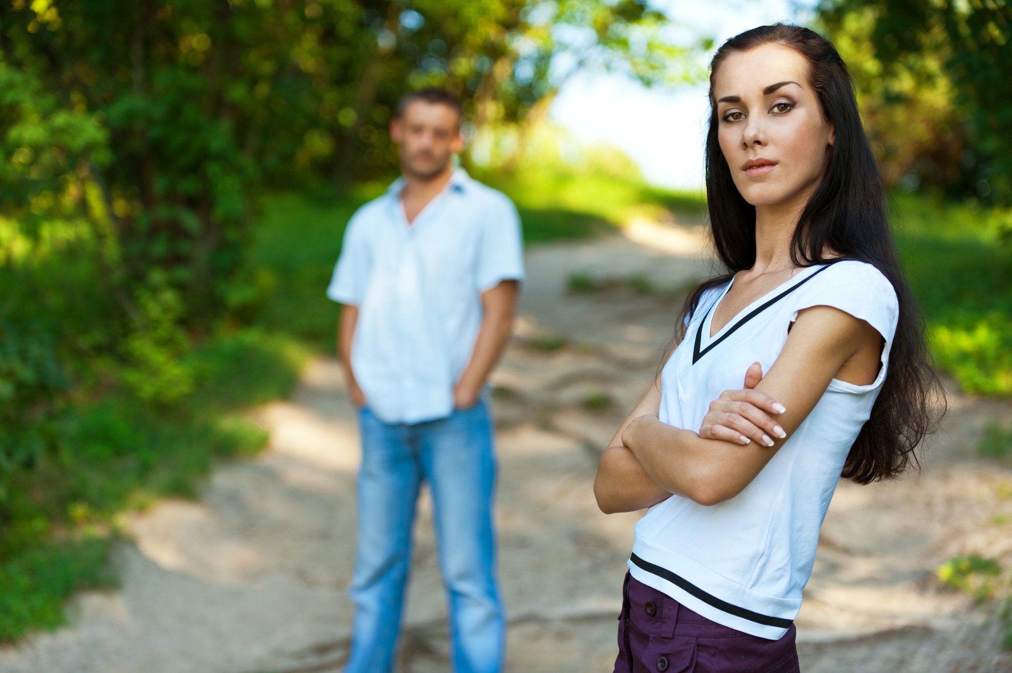 A woman with long dark hair stands in the foreground with arms crossed, wearing a white shirt, and looking at the camera. A man in a white shirt and blue jeans stands blurred in the background on a path surrounded by greenery.