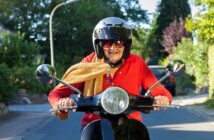 An elderly person wearing a black helmet and sunglasses rides a scooter on a sunny day. They are dressed in a red sweater and have a beige scarf. The background features trees and a street.