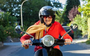 An elderly person wearing a black helmet and sunglasses rides a scooter on a sunny day. They are dressed in a red sweater and have a beige scarf. The background features trees and a street.