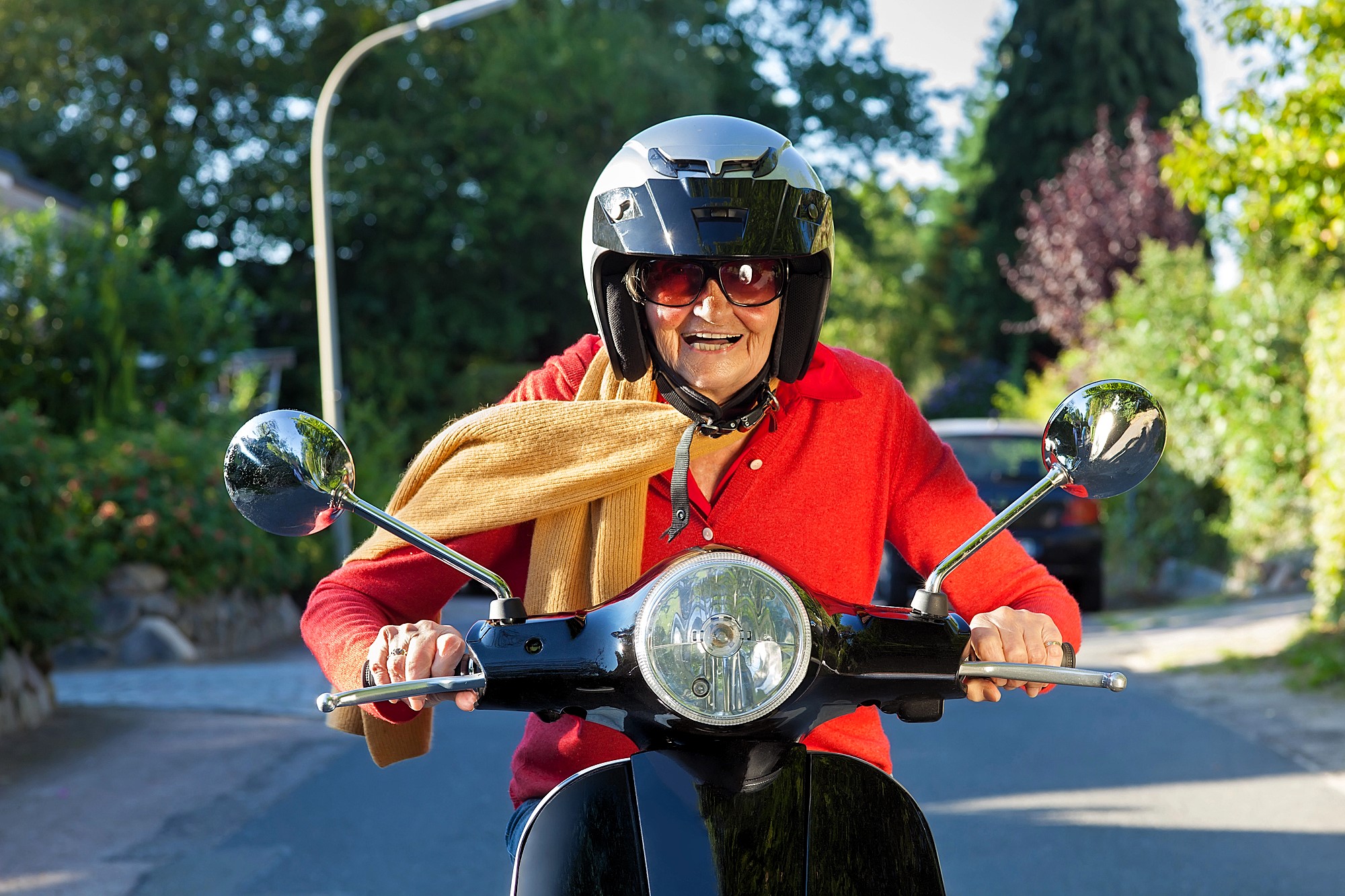 An elderly person wearing a black helmet and sunglasses rides a scooter on a sunny day. They are dressed in a red sweater and have a beige scarf. The background features trees and a street.