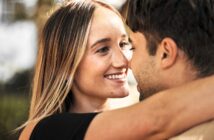 A couple embracing closely, smiling at each other. The woman has long, straight hair, while the man's face is partially visible. The background is blurred, suggesting an outdoor setting.