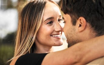 A couple embracing closely, smiling at each other. The woman has long, straight hair, while the man's face is partially visible. The background is blurred, suggesting an outdoor setting.