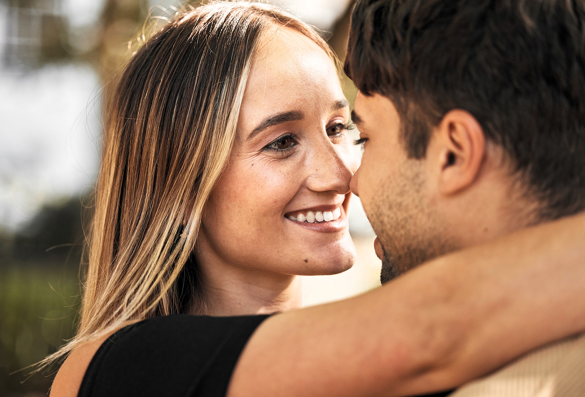 A couple embracing closely, smiling at each other. The woman has long, straight hair, while the man's face is partially visible. The background is blurred, suggesting an outdoor setting.