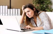 A woman sits at a desk, looking frustrated as she works on her laptop. She rests her head on her hand and leans forward, appearing focused and concerned. In the background, there are shelves with binders and a potted plant.