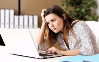 A woman sits at a desk, looking frustrated as she works on her laptop. She rests her head on her hand and leans forward, appearing focused and concerned. In the background, there are shelves with binders and a potted plant.