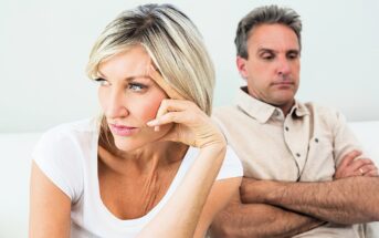 A woman and a man sitting on a couch, both appearing upset. The woman, wearing a white shirt, looks away with a thoughtful expression, resting her hand on her forehead. The man, in a beige shirt, sits with his arms crossed, looking serious.