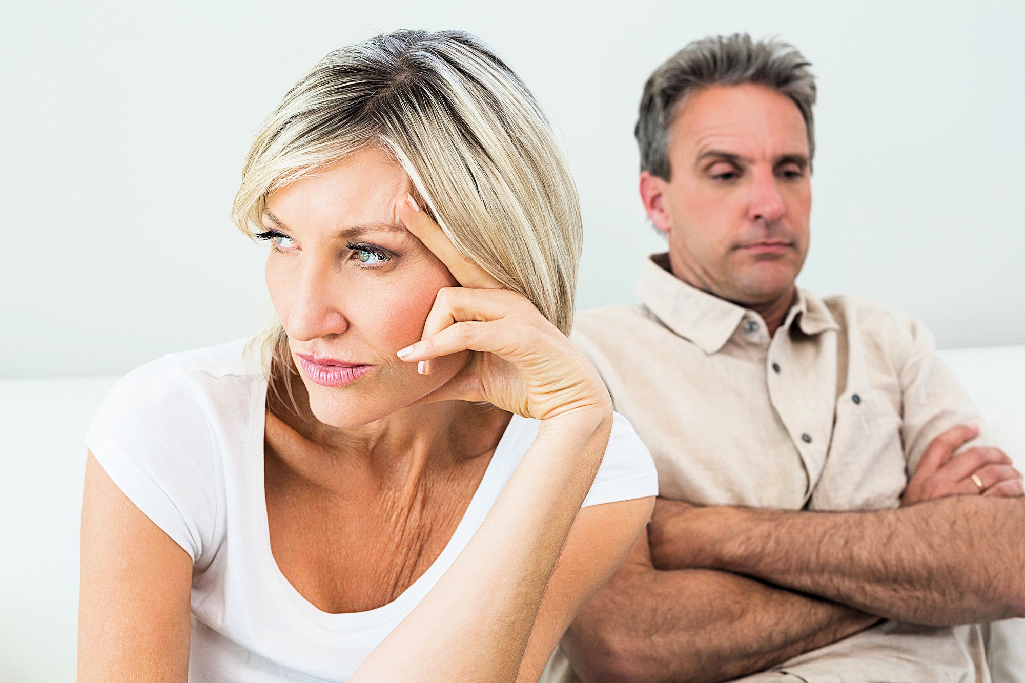 A woman and a man sitting on a couch, both appearing upset. The woman, wearing a white shirt, looks away with a thoughtful expression, resting her hand on her forehead. The man, in a beige shirt, sits with his arms crossed, looking serious.