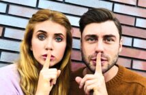 A woman and a man stand side by side in front of a brick wall, each holding a finger to their lips in a gesture suggesting silence. They look directly at the camera. The woman has long blonde hair, and the man has dark hair and a beard.