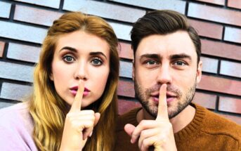 A woman and a man stand side by side in front of a brick wall, each holding a finger to their lips in a gesture suggesting silence. They look directly at the camera. The woman has long blonde hair, and the man has dark hair and a beard.