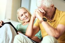 An elderly woman with light hair, seated on a couch, holds a tablet and looks concernedly at an elderly man sitting beside her with his head in his hands, wearing a yellow shirt. The setting appears to be a living room.