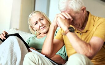 An elderly woman with light hair, seated on a couch, holds a tablet and looks concernedly at an elderly man sitting beside her with his head in his hands, wearing a yellow shirt. The setting appears to be a living room.