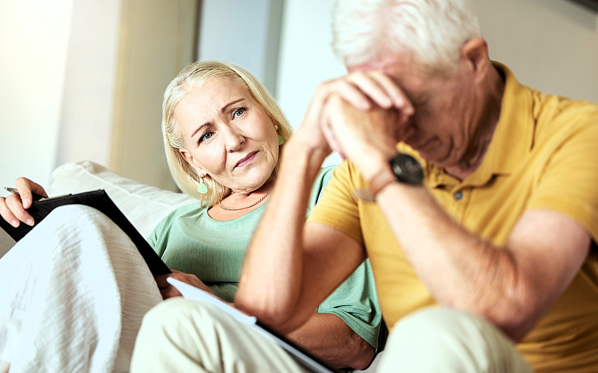 An elderly woman with light hair, seated on a couch, holds a tablet and looks concernedly at an elderly man sitting beside her with his head in his hands, wearing a yellow shirt. The setting appears to be a living room.