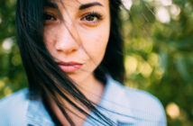 Close-up of a person with long dark hair that is slightly windswept, looking directly at the camera. They are wearing a blue and white striped shirt. The background is a soft focus of greenery.