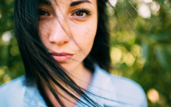 Close-up of a person with long dark hair that is slightly windswept, looking directly at the camera. They are wearing a blue and white striped shirt. The background is a soft focus of greenery.