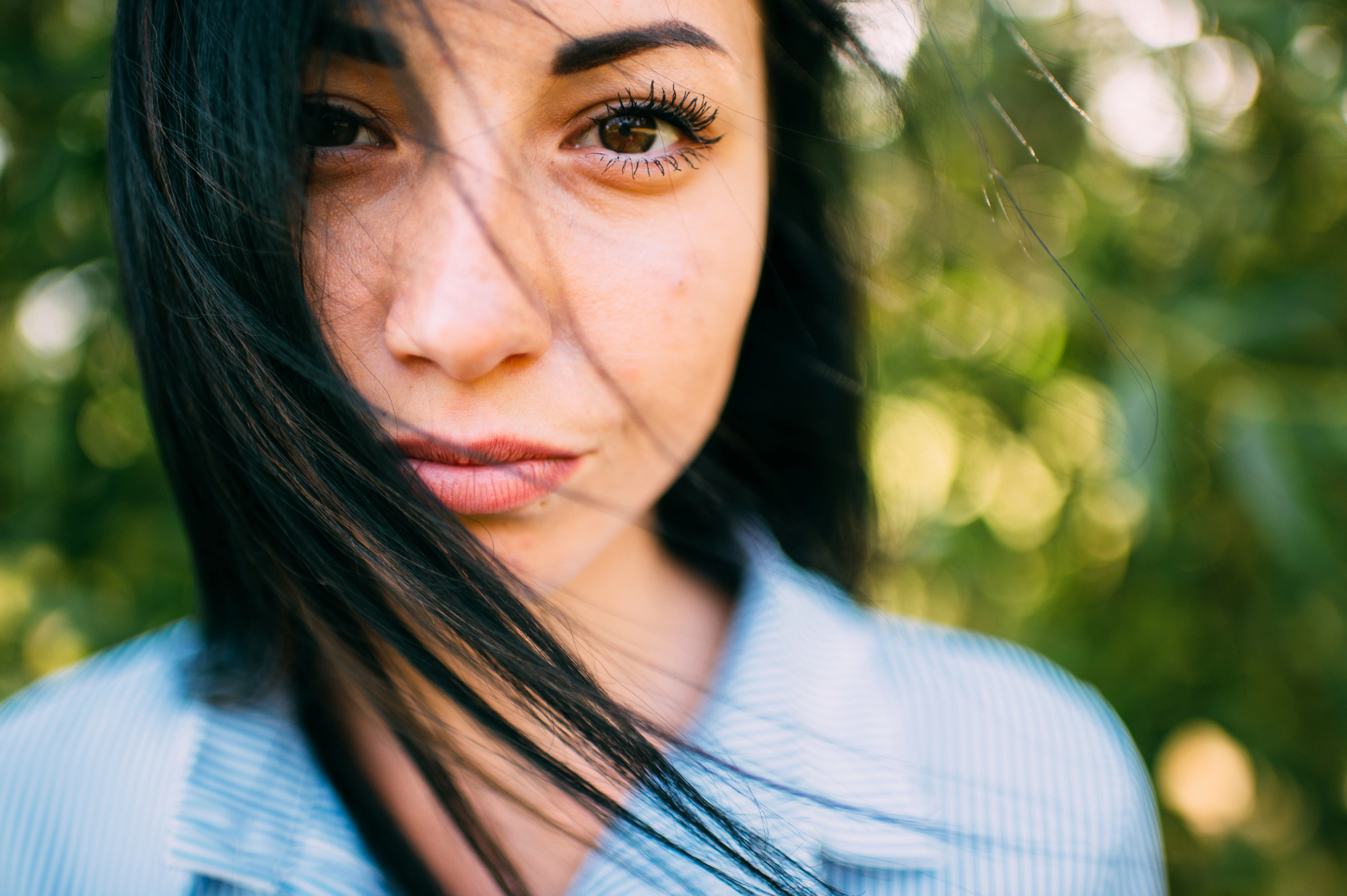 Close-up of a person with long dark hair that is slightly windswept, looking directly at the camera. They are wearing a blue and white striped shirt. The background is a soft focus of greenery.