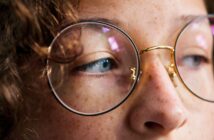 Close-up of a person's face wearing round glasses. The focus is on their eye and the bridge of the glasses, with curly hair partially visible. The lighting highlights the texture of their skin and the glass lenses.