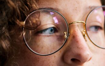 Close-up of a person's face wearing round glasses. The focus is on their eye and the bridge of the glasses, with curly hair partially visible. The lighting highlights the texture of their skin and the glass lenses.