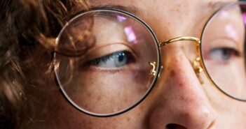 Close-up of a person's face wearing round glasses. The focus is on their eye and the bridge of the glasses, with curly hair partially visible. The lighting highlights the texture of their skin and the glass lenses.