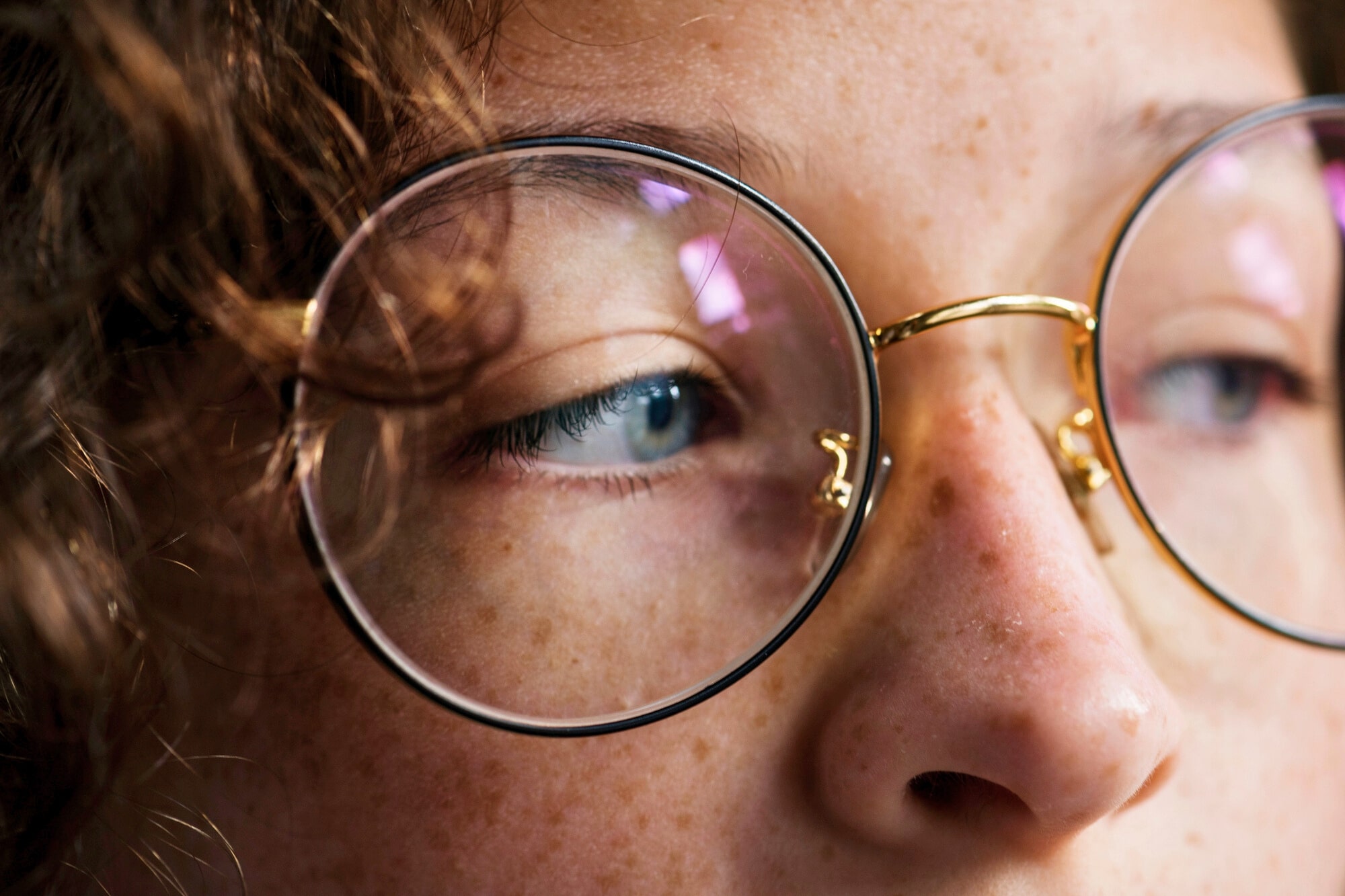 Close-up of a person's face wearing round glasses. The focus is on their eye and the bridge of the glasses, with curly hair partially visible. The lighting highlights the texture of their skin and the glass lenses.