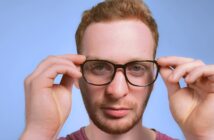 A person with short, reddish-brown hair and a beard is looking at the camera, adjusting black-rimmed glasses with both hands. The background is a plain blue.