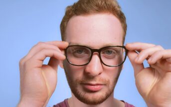 A person with short, reddish-brown hair and a beard is looking at the camera, adjusting black-rimmed glasses with both hands. The background is a plain blue.