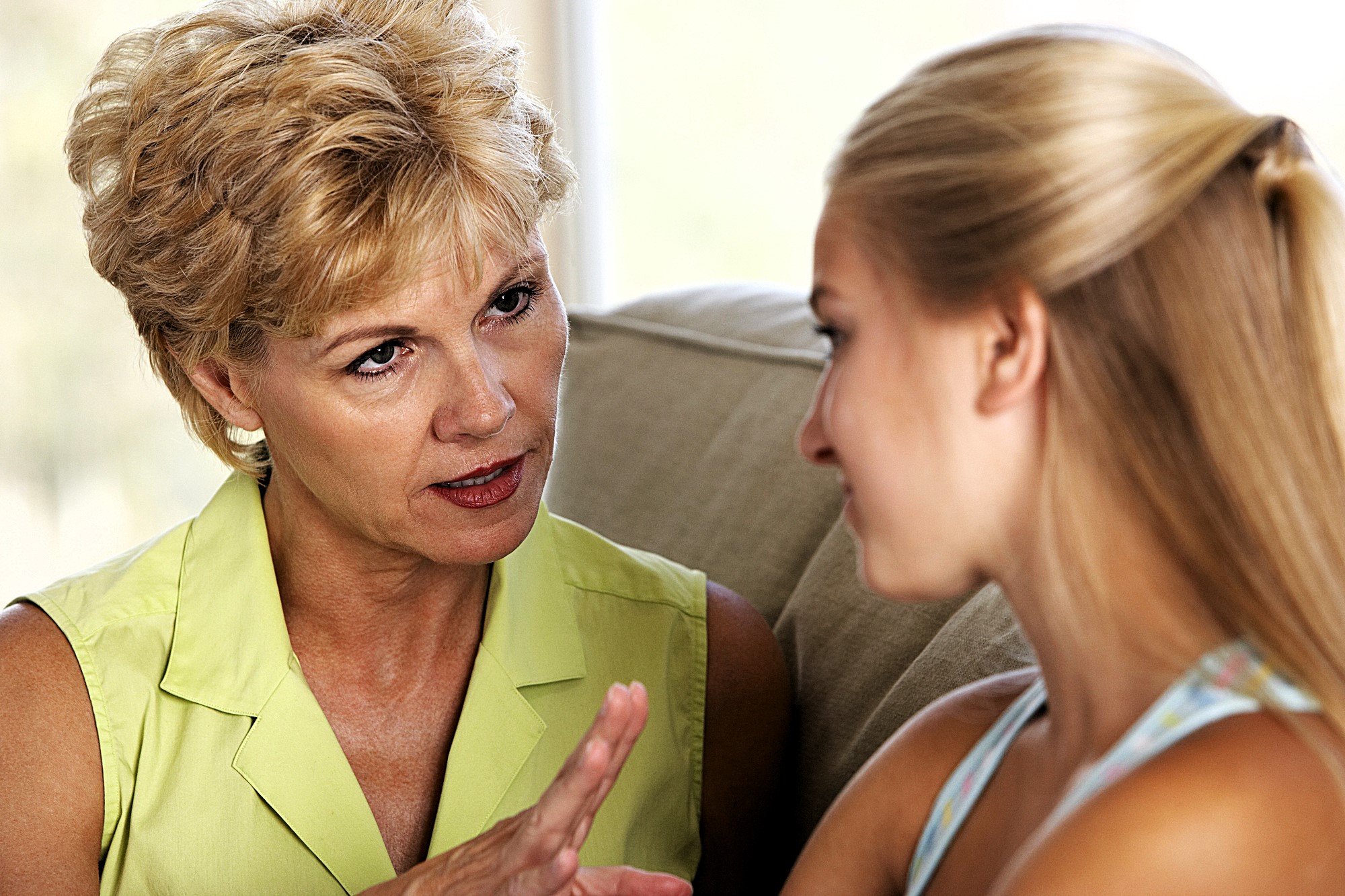 A woman with short blond hair is speaking seriously to a younger woman with long blond hair. They are sitting on a couch indoors, and the older woman is gesturing with her hand.