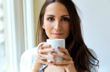 A woman with long brown hair is standing by a window, holding a white mug with both hands. She is looking directly at the camera with a relaxed, neutral expression. The background is softly lit by natural light.