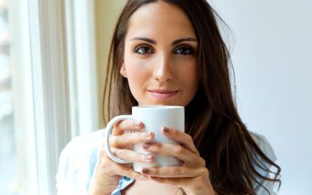 A woman with long brown hair is standing by a window, holding a white mug with both hands. She is looking directly at the camera with a relaxed, neutral expression. The background is softly lit by natural light.