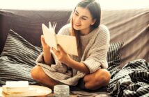 A woman sits cross-legged on a couch, wearing a cozy sweater, and reads a book with a smile. A wooden tray with a mug and an open book is beside her. The setting appears relaxed and comfortable.