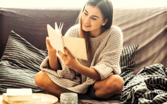 A woman sits cross-legged on a couch, wearing a cozy sweater, and reads a book with a smile. A wooden tray with a mug and an open book is beside her. The setting appears relaxed and comfortable.