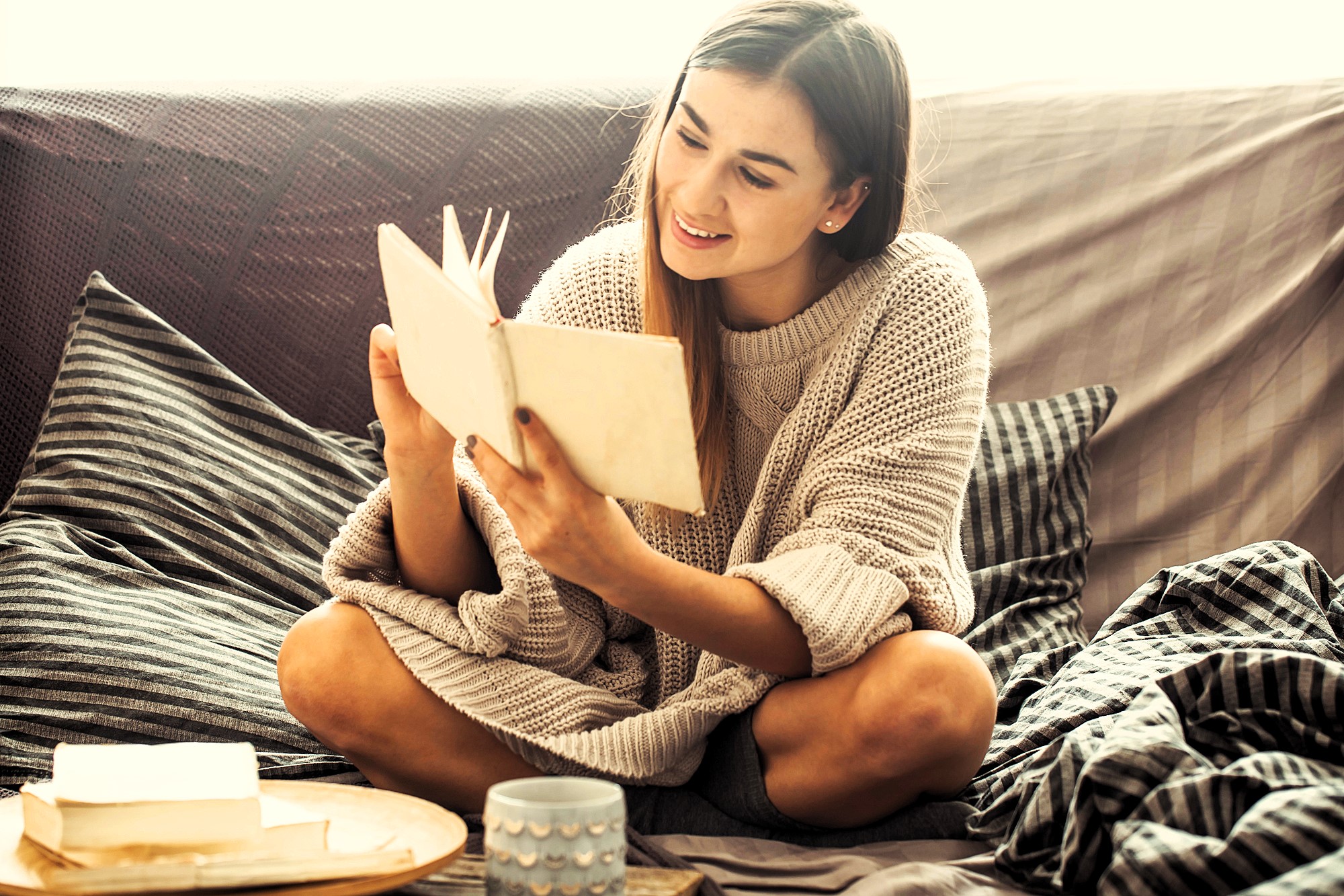 A woman sits cross-legged on a couch, wearing a cozy sweater, and reads a book with a smile. A wooden tray with a mug and an open book is beside her. The setting appears relaxed and comfortable.