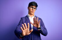 A man with black hair and glasses stands in front of a purple background. He is wearing a blue blazer and a white shirt, extending his hands forward with a cautious or defensive expression.