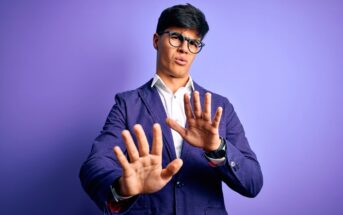 A man with black hair and glasses stands in front of a purple background. He is wearing a blue blazer and a white shirt, extending his hands forward with a cautious or defensive expression.