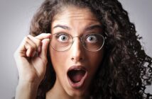 A person with curly hair, wearing round glasses, has an expression of surprise with mouth open and eyes wide. The hand is adjusting the glasses. The background is plain and light-colored.