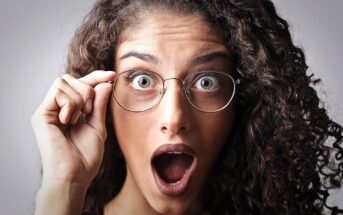 A person with curly hair, wearing round glasses, has an expression of surprise with mouth open and eyes wide. The hand is adjusting the glasses. The background is plain and light-colored.