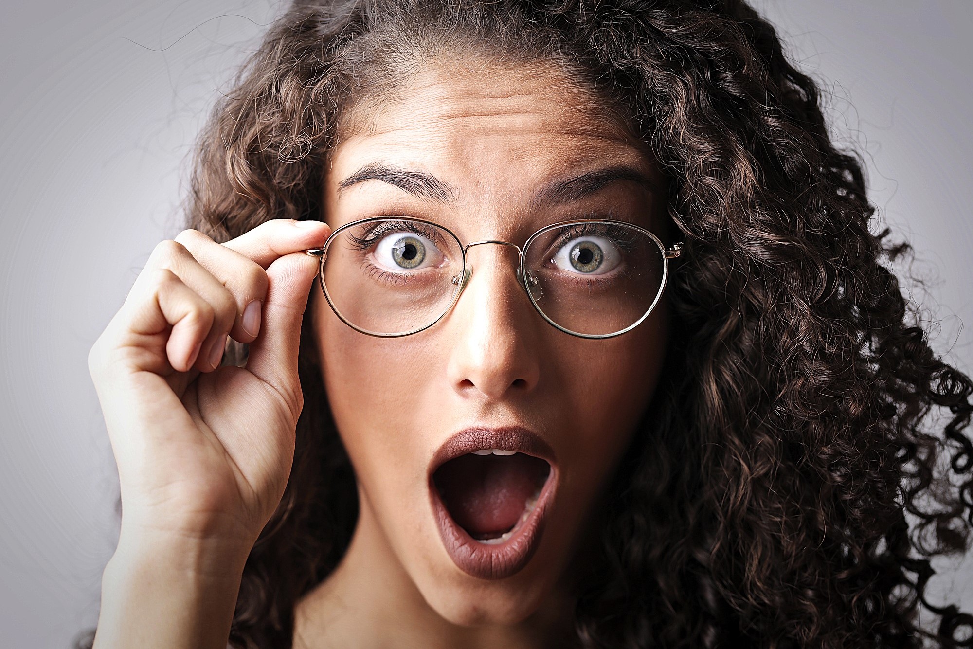 A person with curly hair, wearing round glasses, has an expression of surprise with mouth open and eyes wide. The hand is adjusting the glasses. The background is plain and light-colored.