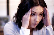 A woman with dark hair holds her hands to her temples, appearing stressed or deep in thought. She is wearing a light-colored sweater and looking slightly downward. The background is softly blurred.